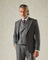 A man wearing a grey pinstripe suit jacket with a single-breasted, single-button closure, peak lapel, horn buttons, and a double vent. The jacket features a subtle striped pattern with Morse code stitches.